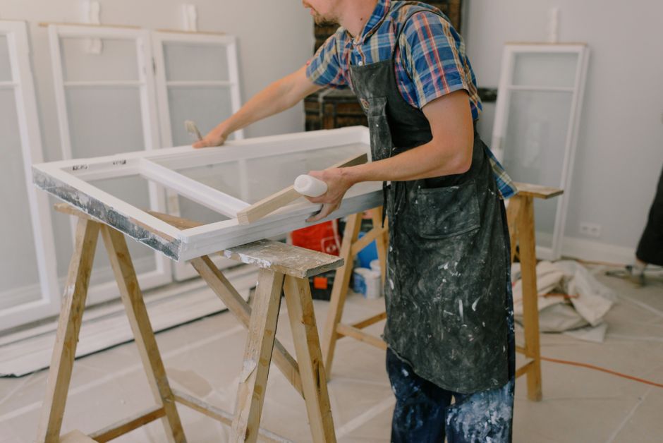 A craftsman standing near a white window frame on a workbench.